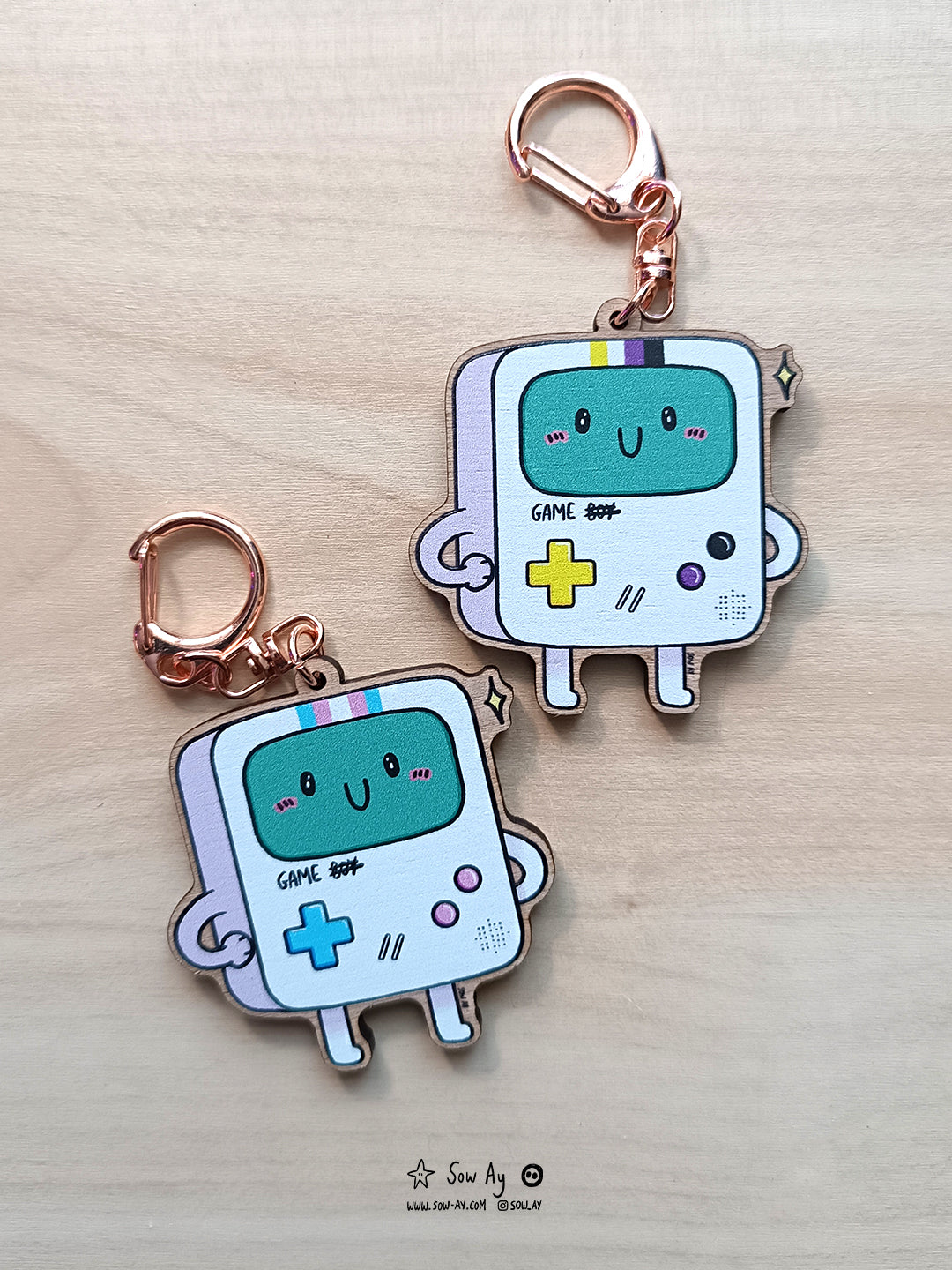 une petite console de jeu portable, aux couleurs du drapeau transgenre ou non binaire. Il est écrit game boy et le boy est raturé.