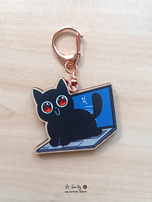 Petit chat noir avec un grand sourire, de grands yeux aux pupilles très dilatées dans lesquelles on voit des flammes. Il est allongé sur le clavier d'un ordinateur portable. Sur l'écran du pc, on voit du bleu, relatif aux écrans de bug avec un emoji pas content.