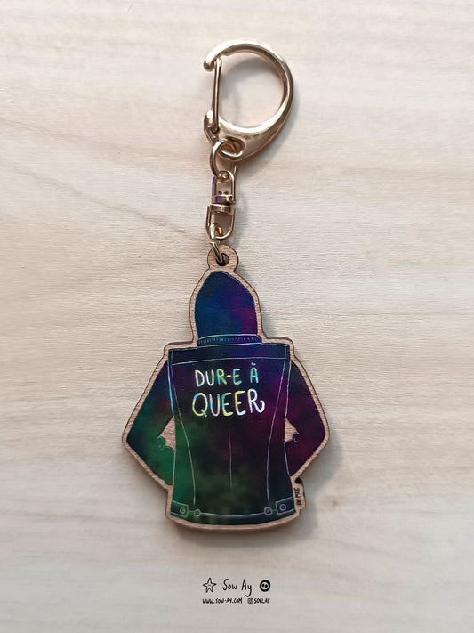 Personnage de dos jusqu'à la taille : il porte une veste noire et il y a écrit dessus en multicolore : dur à queer