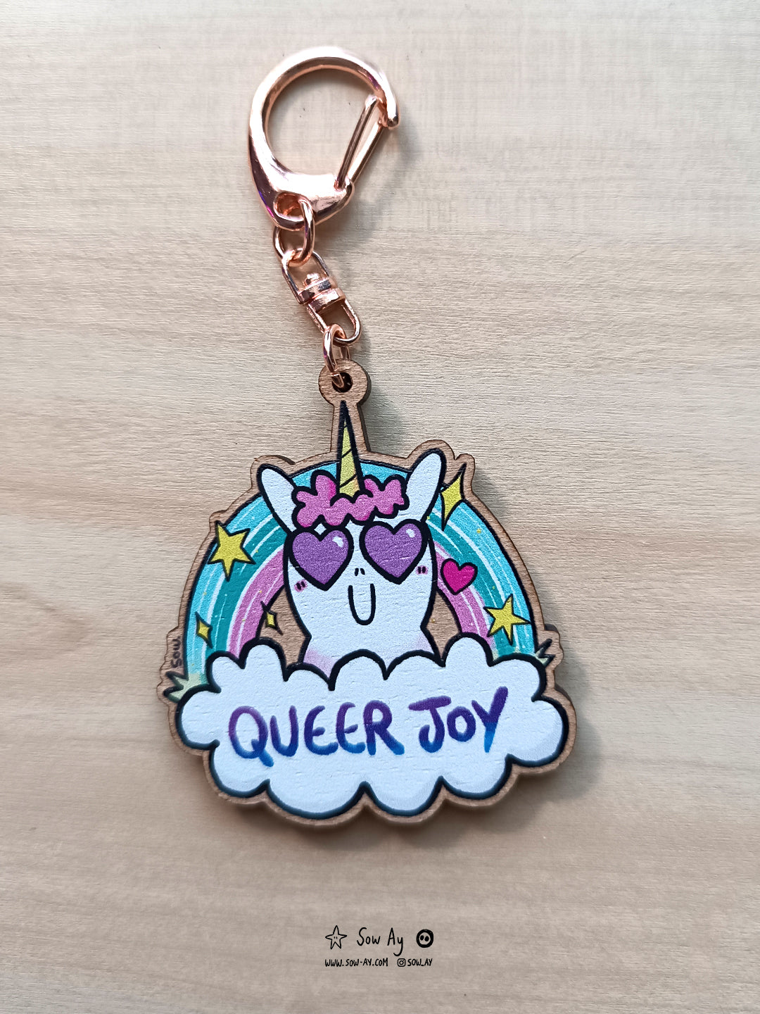 une petite licorne avec un grand sourire et deux coeurs dans les yeux. Derrière elle, un arc en ciel aux couleurs turquoises et roses. Devant elle, un nuage dans lequel est écrit en violet "queer joy".