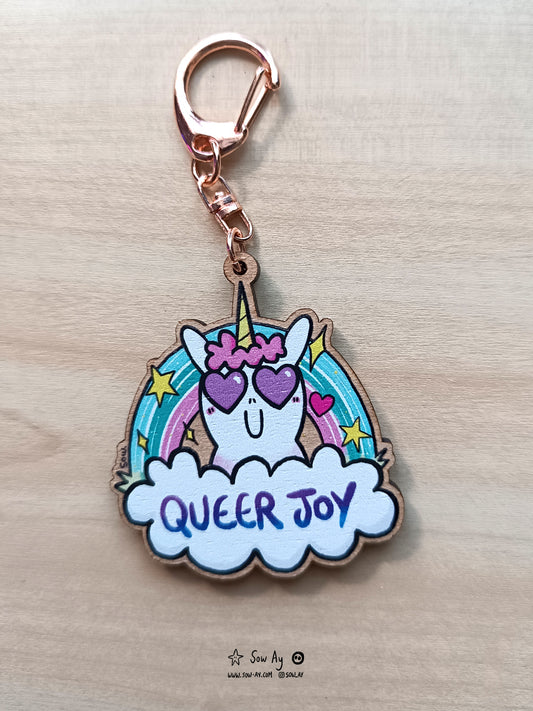 une petite licorne avec un grand sourire et deux coeurs dans les yeux. Derrière elle, un arc en ciel aux couleurs turquoises et roses. Devant elle, un nuage dans lequel est écrit en violet "queer joy".
