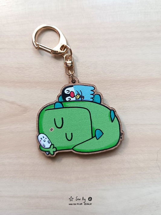 Petit dinosaure vert qui dort en boule, il tient dans sa main un doudou de fantôme et sur son dos, dort son ami pingouin.