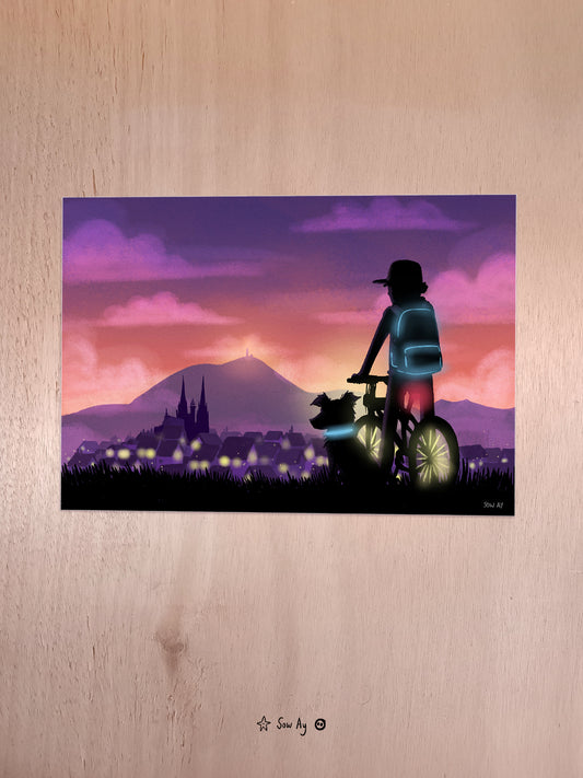 le jour se lève, il fait encore bien nuit, on voit de dos un garçon avec une casquette et un sac à dos sur son vélo. A côté, son chien est assis, également de dos. Au loin, on voit la ville de Clermont-Ferrand et le Puy-de-dôme.