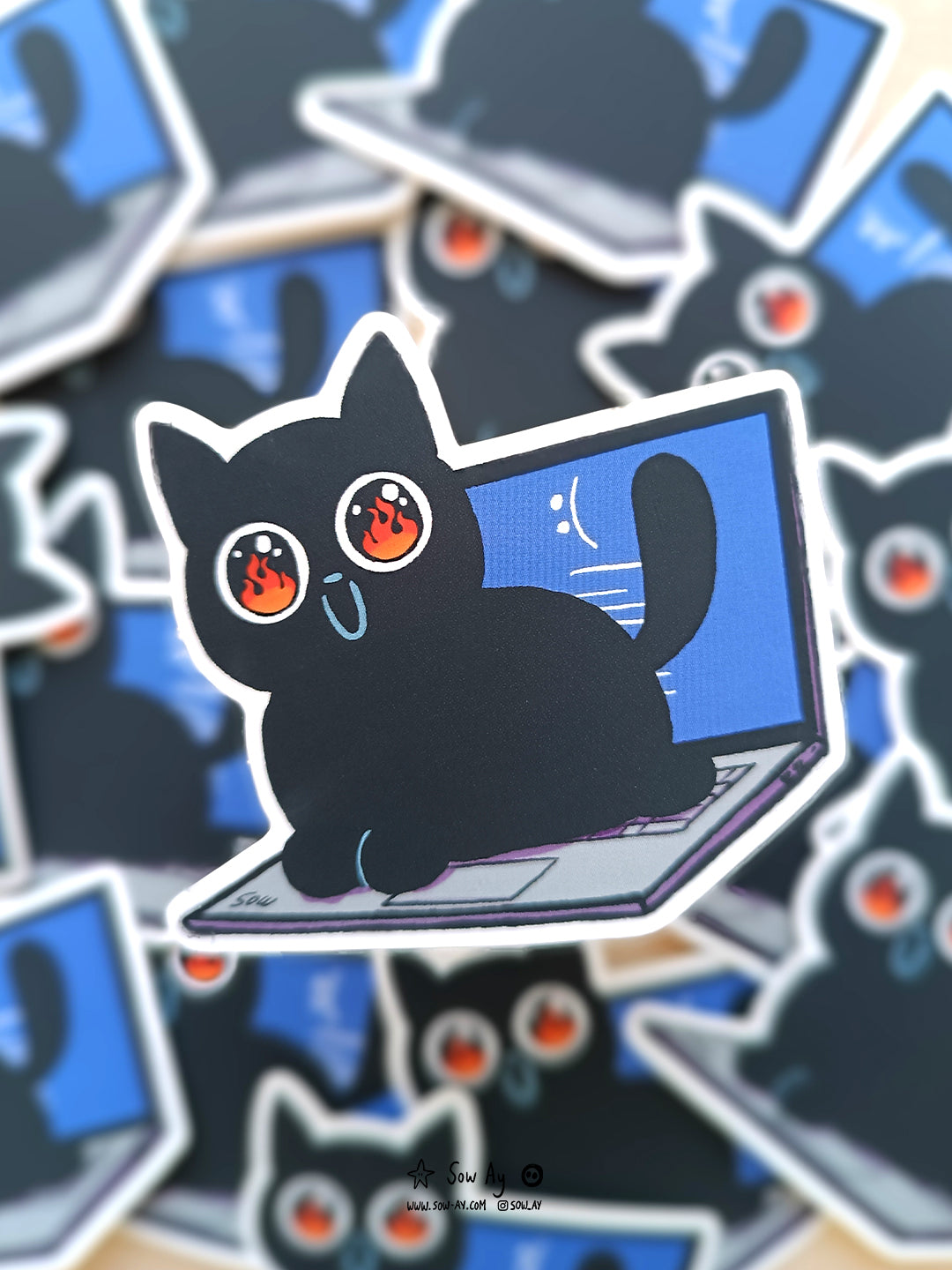chat noir au grand sourire et immenses yeux aux pupilles dilatées dans lesquelles on voit des flammes. il est assis sur le clavier d'un ordinateur portable, sur lequel on aperçoit un écran bleu, signe de bug.