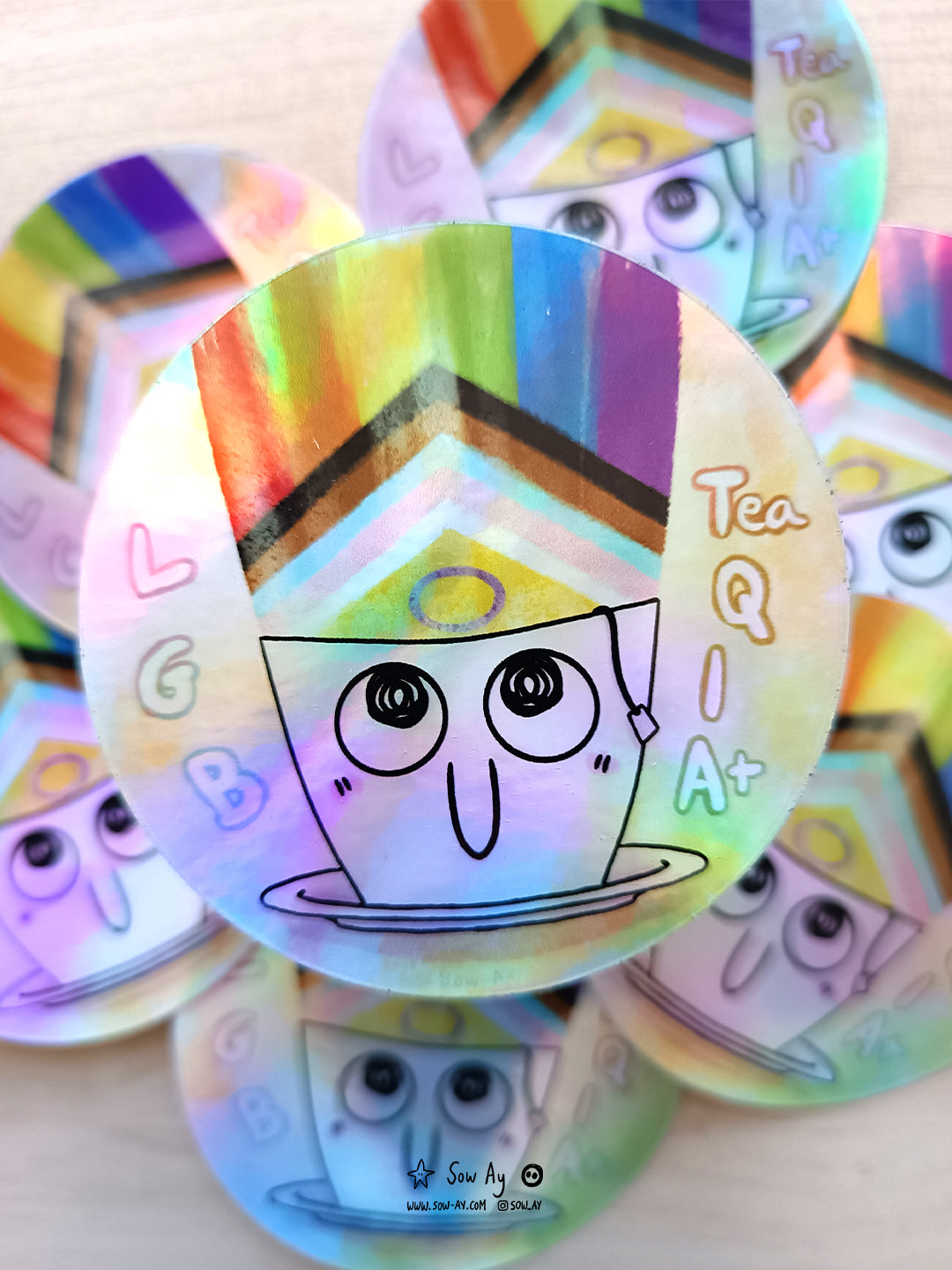 une tasse blanche avec le bout du sachet de thé qui dépasse. Elle sourit et le drapeau LGBTQIA sort vers le haut. Il est écrit le jeu de mot LGB tea QIA+