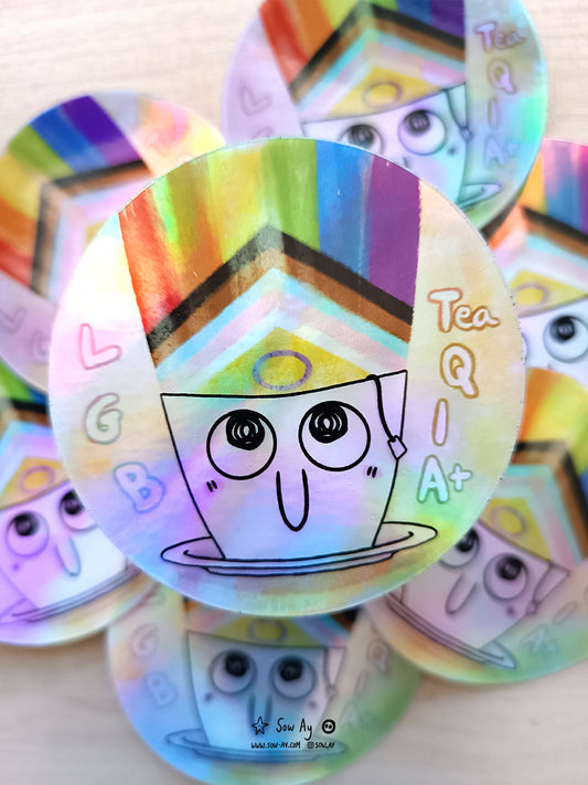 une tasse blanche avec le bout du sachet de thé qui dépasse. Elle sourit et le drapeau LGBTQIA sort vers le haut. Il est écrit le jeu de mot LGB tea QIA+