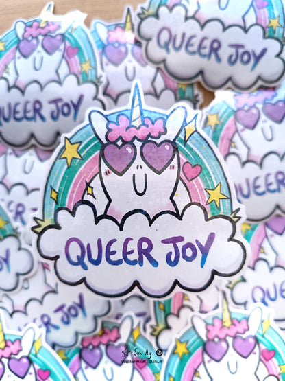une tête licorne de face avec des cœurs dans ses yeux. Il y a un arc en ciel derrière, et devant elle un nuage dans lequel il y est écrit "queer joy"