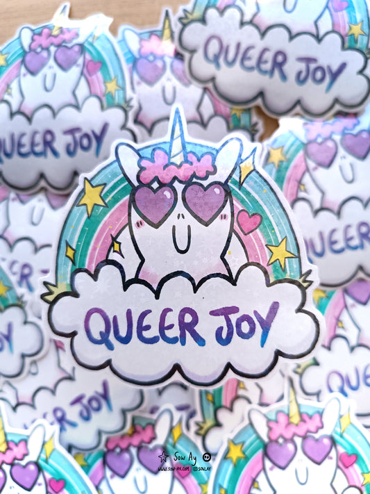 une tête licorne de face avec des cœurs dans ses yeux. Il y a un arc en ciel derrière, et devant elle un nuage dans lequel il y est écrit "queer joy"